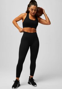 Schwarzer Sport-BH mit Rundhalsausschnitt, kombiniert mit hochgeschnittenen schwarzen Leggings. Das Model trägt schwarze Sportschuhe und hält einen braunen Basketball.