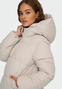 Giacca a vento in beige chiaro, con un ampio cappuccio, design trapuntato e chiusura con zip frontale. Tessuto morbido con tasche laterali.