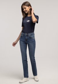 Granatowy bawełniany t-shirt z białym logo, w połączeniu z jasnoniebieskimi dżinsami i białymi trampkami, w pozycji stojącej na neutralnym tle.