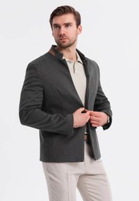 Blazer gris con un cuello alto y detalles marrones, abotonado. Llevado sobre una camiseta polo beige, combinado con pantalones de color claro. Textura suave.