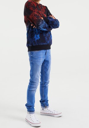 Neulepuserossa on punaisia ja sinisiä trooppisia kukkakuvioita, mustat hihansuut ja helma. Yhdistetty vaaleansinisiin farkkuihin ja valkoisiin lenkkareihin, joissa on punaisia yksityiskohtia.