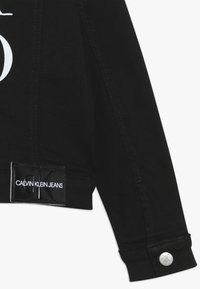 Schwarze cropped Jacke aus Baumwollmischung. Verfügt über ein weißes Logo und silberne Knopfdetails an den Manschetten. Glatte Textur mit Nähakzenten.