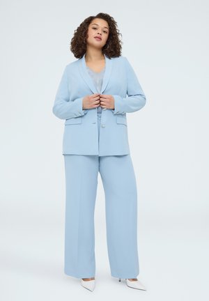 Kvinde med krøllet hår iført en lyseblå dragt med bukser med brede ben og hvide spidse hæle, stående mod en ensfarvet hvid baggrund.