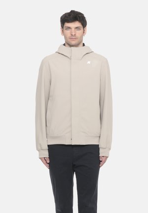 Giacca beige con cappuccio e chiusura a zip, caratterizzata da una texture liscia, polsini a coste e un logo discreto sul petto. Pantaloni neri completano il look.