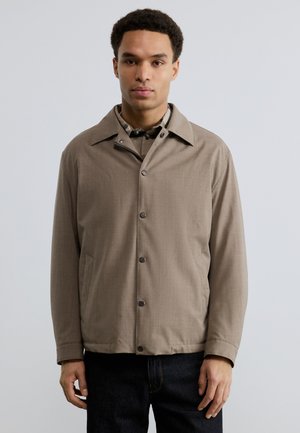 Νεαρός άνδρας που φοράει μπεζ σακάκι με κουμπιά πάνω από ένα καρό πουκάμισο, στέκεται μπροστά σε μια απλή ανοιχτόχρωμη επιφάνεια.