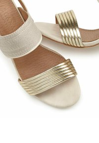 Beige sandalen met een getextureerde tan binnenzool, voorzien van een gestreepte witte band en een glanzende gouden band met horizontale groeven.