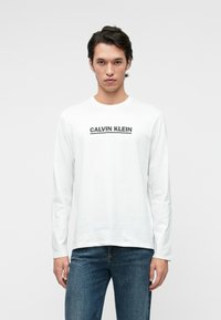 Dolgimi rokavi bela bombažna majica s črnim logotipom "CALVIN KLEIN" čez prsa, v kombinaciji z modrimi kavbojkami, preprosta ozadje.