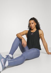 Črna brezrokavna majica s majhnim roza Nike logotipom, kombinirana z svetlo modrimi leginsami z teksturiranimi detajli in sivi športni čevlji.