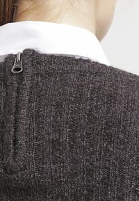 Harmaa, struktuurillinen neulepaita, jossa on pystysuorat resorit ja selässä näkyvä metallivetoketju, yhdistetty valkoiseen kauluspaitaan.