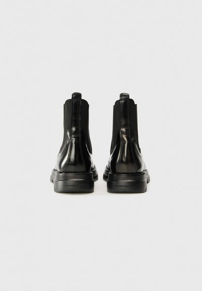 Bottes Chelsea noires à la texture lisse et brillante, avec des panneaux latéraux élastiques et une semelle épaisse noire, comportant un logo discret sur le talon.