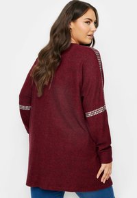 Burgundyröd långärmad tröja med avslappnad passform, som har glänsande nitar på ärmarna och ett mjukt, texturerat tyg.