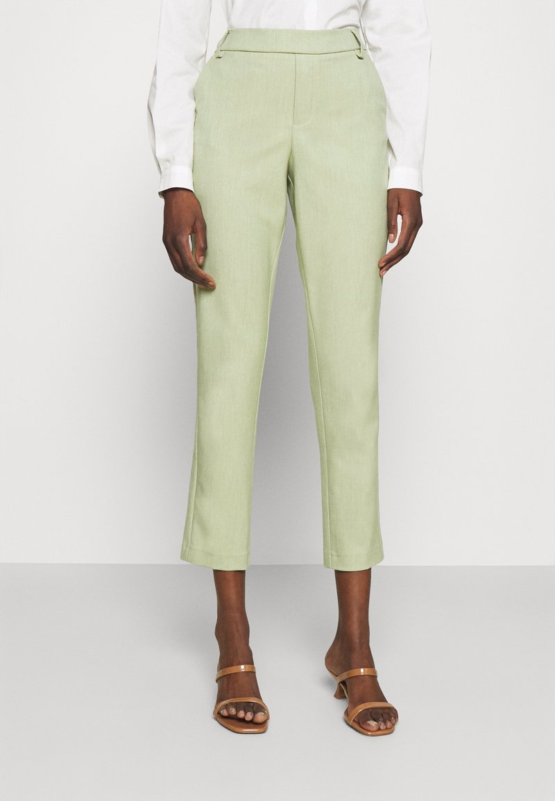 Pantalon vert clair ajusté avec une texture lisse, coupe slim et longueur raccourcie ; associé à des sandales marron à brides et une chemise blanche à manches longues.