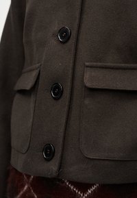 Σκούρο καφέ μάλλινο σακάκι με μεγάλα μαύρα κουμπιά και δύο τσέπες με καπάκι, φορεμένο πάνω από μια καφέ φούστα με σχέδιο.