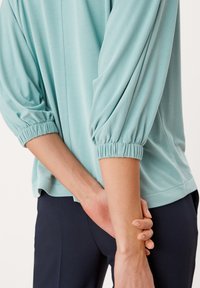 Top azzurro chiaro con maniche a palloncino e polsini elastici, realizzato in un tessuto morbido. Il design presenta una vestibilità rilassata e un orlo arrotondato.