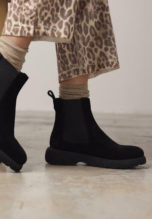Botines de ante negro con paneles elásticos laterales, suela de goma texturizada y tira de sujeción. Combinados con calcetines de punto beige y pantalones de leopardo.