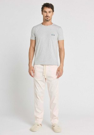 Homme debout, portant un t-shirt gris clair avec un petit logo de bicyclette sur la poitrine, un pantalon ample beige clair et des chaussures beige enfilables, sur un fond uni.