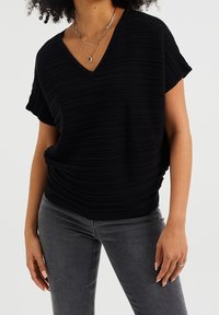 Černý pletený top s výstřihem do V a krátkými rukávy, který má strukturovaný, proužkovaný design. V kombinaci se šedými džíny, které podtrhují ležérní vzhled.