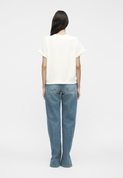 Γυναίκα με μακριά σκούρα μαλλιά που φοράει άσπρο φαρδύ κοντομάνικο τοπ και μπλε τζιν με ίσια γραμμή, στέκεται γυρισμένη με την πλάτη προς ένα απλό φόντο.