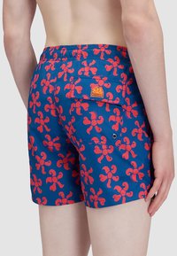 Pantaloni da nuoto blu con un motivo floreale rosso, vita elastica e tasca posteriore con un'etichetta del logo. Il tessuto ha una texture liscia.