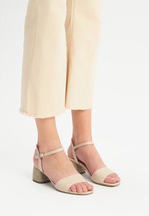 Beżowe sandały na wysokim obcasie z paskowym wzorem, z teksturowanym obcasem i regulowanym paskiem wokół kostki, noszone z jasnymi spodniami 3/4.