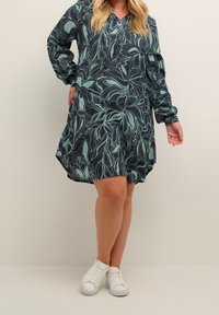 Robe imprimée florale en bleu marine avec un motif de feuilles abstraites vertes, manches longues, décolleté en V et forme évasée. Associée à des baskets blanches.