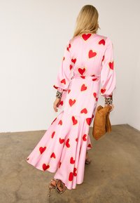 Γυναίκα με μακριά ξανθά μαλλιά που φοράει ένα ροζ φόρεμα με κόκκινες και χρυσές καρδιές, μανσέτες και τακούνια με λεοπάρ σχέδιο, κρατώντας μία καφέ τσάντα από σουέτ.