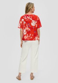 Rotes Blusenshirt mit floralem Druck und kurzen Ärmeln, versehen mit weißen und blauen Akzenten, kombiniert mit weißen weitem Hosen. Glatte Stoffstruktur.