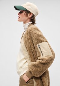 Brūna, pūkaina virsdrēbe ar ielīmētu bezkrāsas datiņu un rāvējslēdzēja kabatu; papildināta ar baltiem augstu apkakli un bēša/zaļu beisbola cepuri.