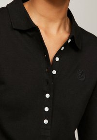 Schwarzes Polohemd aus strukturiertem Stoff mit Kragen, langen Ärmeln und fünf weißen Knöpfen entlang der Knopfleiste sowie einem gestickten Logo.