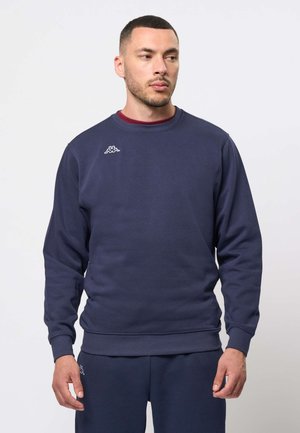 Mann trägt dunkelblauen Sweatshirt und passende Hose mit kleinem weißem Logo, schaut leicht nach links, vor einfachem hellem Hintergrund.