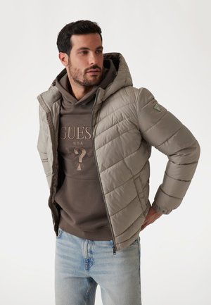 Gevoerde jas in gedempt beige met een grijze capuchon. Heeft een gequilt ontwerp, een rits aan de voorkant en een logo detail op de linkerschouder. Draagt over een donkere hoodie.