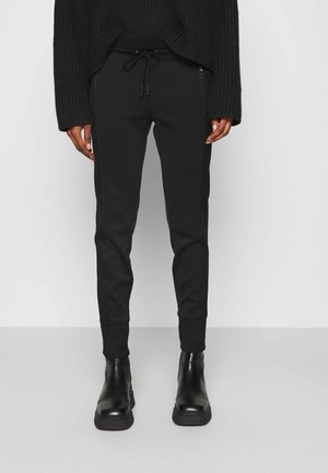 Personne portant un pantalon de jogging noir à cordon, un pull en maille noir et des bottines noires, debout devant un mur clair et uni.