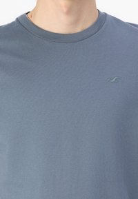 Blå bomulls T-skjorte med rund hals og korte ermer. Har en liten, kontrasterende logo på venstre bryst. Myk, jevn tekstur.
