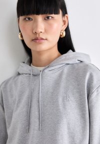 Šedá mikina s kapucňou a šnúrkami, jemná textúra, s nenápadným vyšívaným logom na hrudi. Model má zlaté kruhové náušnice.