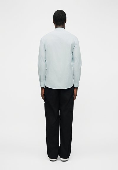 Άνδρας που στέκεται με την πλάτη στην κάμερα, φοράει ανοιχτό μπλε πουκάμισο με μακριά μανίκια, μαύρο παντελόνι και λευκά παπούτσια σε απλό φόντο.