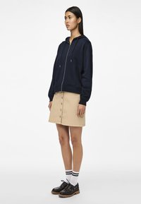 Felpa blu navy con chiusura a zip e tasche a marsupio, abbinata a una gonna beige con bottoni e scarpe nere con suola spessa e calzini bianchi a righe.