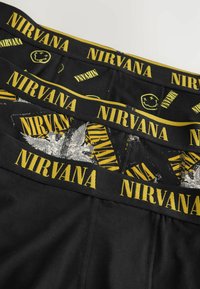 Sous-vêtements noirs avec une ceinture élastique jaune portant le texte "NIRVANA" et un motif de visages souriants et de designs graphiques.