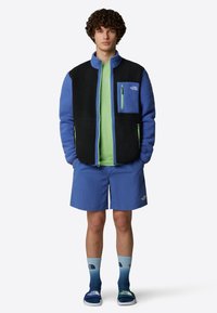 Fleecejacka med svarta och blå sektioner, med en dragkedjeförsedd bröstficka. Bärs över en grön skjorta, tillsammans med blå shorts och graderade strumpor.