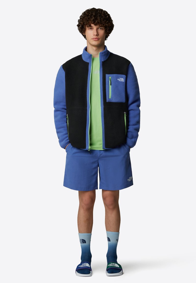 Fleecejacka med svarta och blå sektioner, med en dragkedjeförsedd bröstficka. Bärs över en grön skjorta, tillsammans med blå shorts och graderade strumpor.