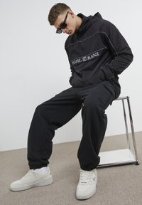 Black lightweight anorak with a hood, featuring a gray logo, paired with black joggers and white high-top sneakers. Smooth fabric texture.