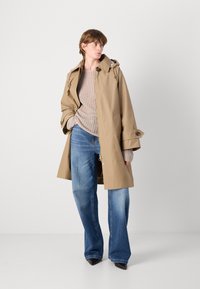 Beige wasserdichter Trenchcoat mit Kapuze, doppelter Knopfleiste und weiten Ärmeln, kombiniert mit einem beigen Strickpullover und blauen Schlagjeans.