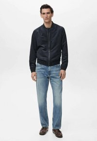 Chaqueta bomber de ante azul marino con puños acanalados, combinada con jeans de pierna recta azul claros y zapatos marrones. Diseño sencillo y moderno con cierre de cremallera.
