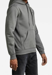 Sudadera gris hecha de tejido suave, con un bolsillo frontal, capucha ajustable y puños de canalé. Textura suave con detalles de costura discretos.
