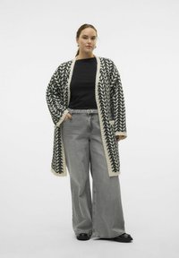 Gilet en tricot avec un motif en chevrons noir et blanc, ouvert à l'avant, et bords crème. Associé à un jean large gris.