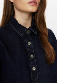 Donkerdenim overhemd met kraag, voorzien van zilveren knoopsluitingen en subtiele contrasterende stiksel. De stof heeft een soepele textuur.