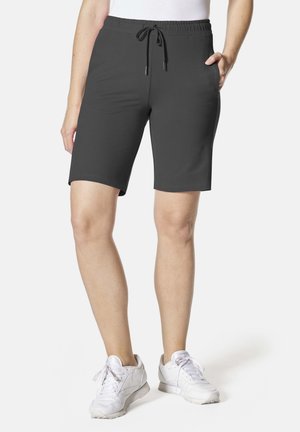Frau trägt schwarze, kniehohe Shorts mit elastischem Bund und Kordelzug sowie weiße Turnschuhe, steht mit einer Hand in der Tasche.
