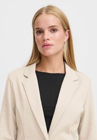 Beige blazer met een gesneden revers, zachte textuur en lichte drape, gedragen over een geribbeld zwart topje. Eenvoudig ontwerp, geen zichtbare hardware of patronen.