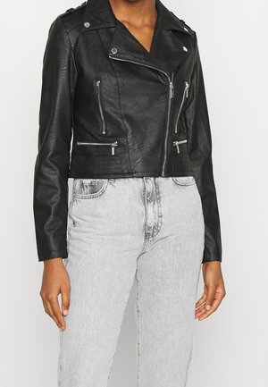 Personne portant une veste courte de biker en similicuir noir avec des fermetures éclair argentées et un jean taille haute gris clair.