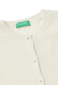 Krémový kardigan vyrobený z měkké pleteniny, s kulatým výstřihem, pěti perlami a zelenou cedulkou značky uvnitř výstřihu.