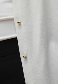 Cardigan bianco con un tessuto a maglia testurizzato, caratterizzato da un elemento decorativo in oro. Il cardigan è indossato sopra un outfit nero.